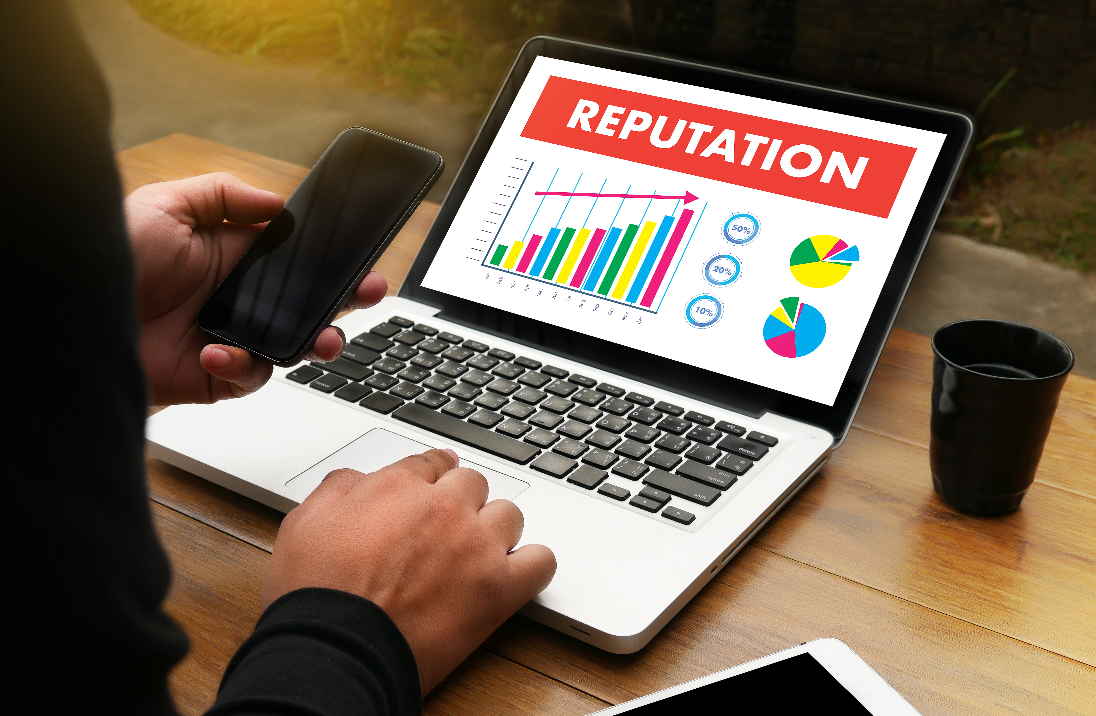 Digital marketer analyzing online reputation management data on a laptop displaying colorful charts, holding a smartphone, with a tablet and coffee cup nearby on a wooden table.