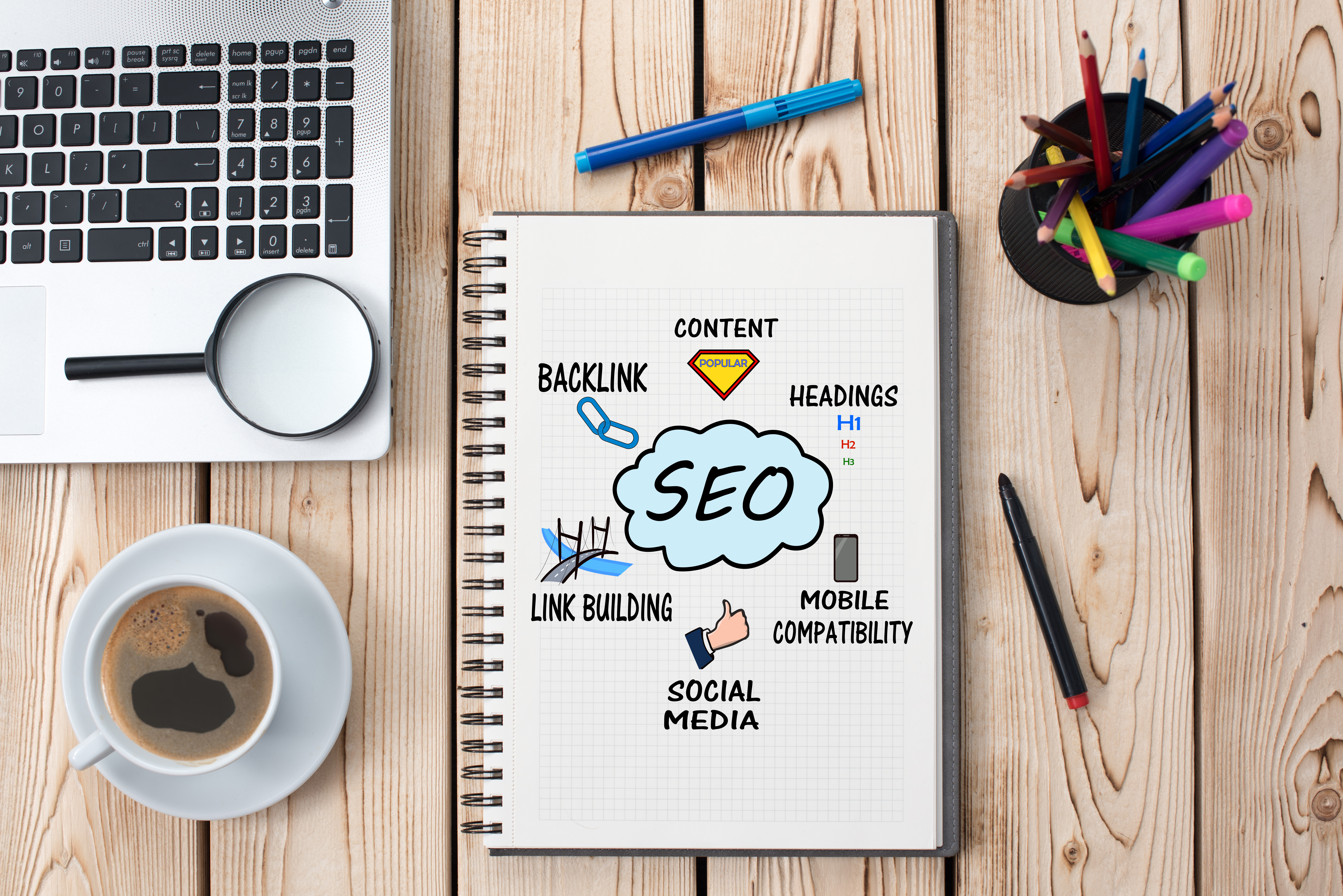 A wooden desktop in the digital marketing agency with an SEO solutions doodle on a notepad, a laptop, colorful pens, a magnifying glass, and a coffee cup.