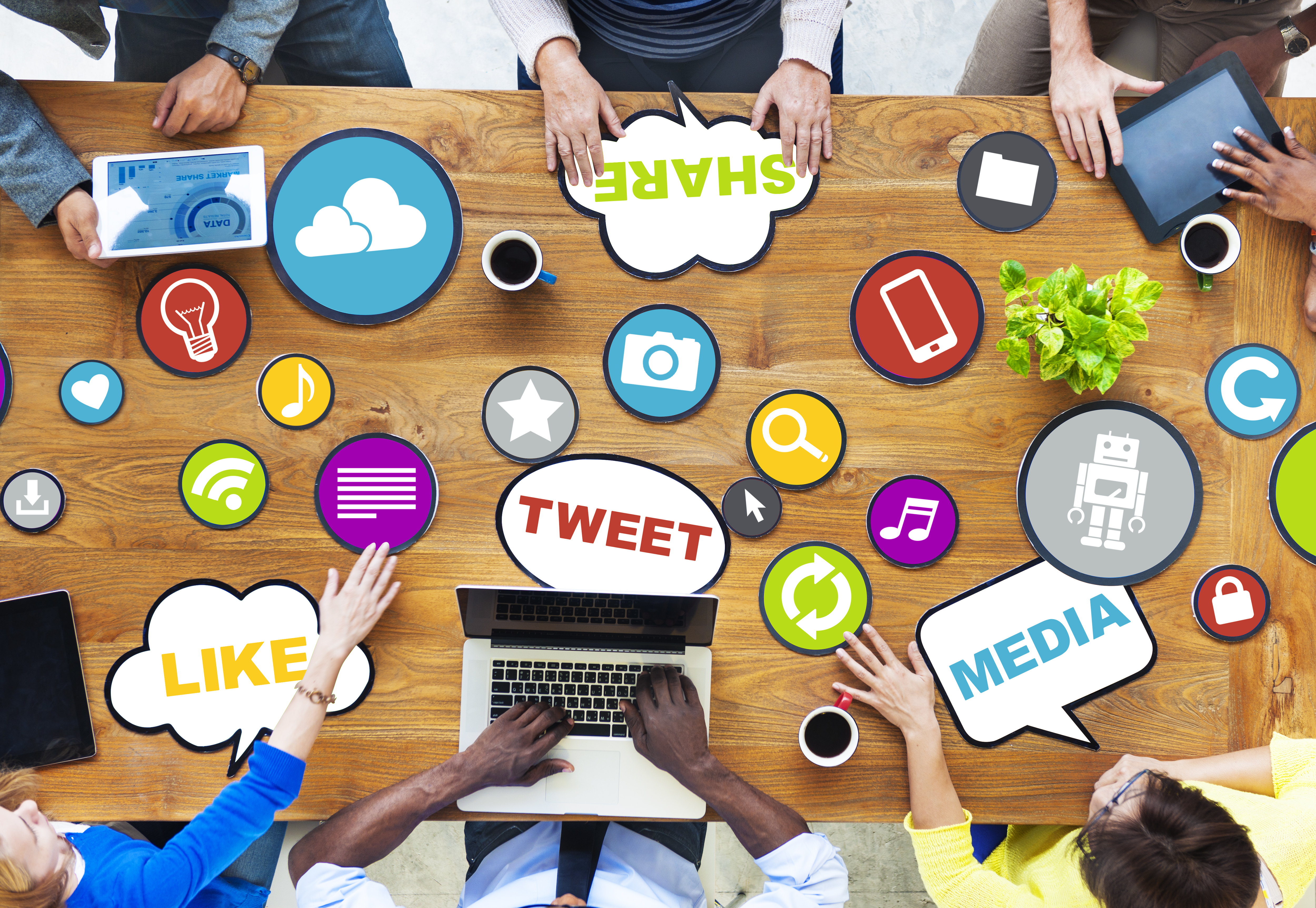 Overhead view of digital marketing team in a brainstorming session with devices, surrounded by social media icons including 'like', 'share', 'tweet', on a wooden table.
